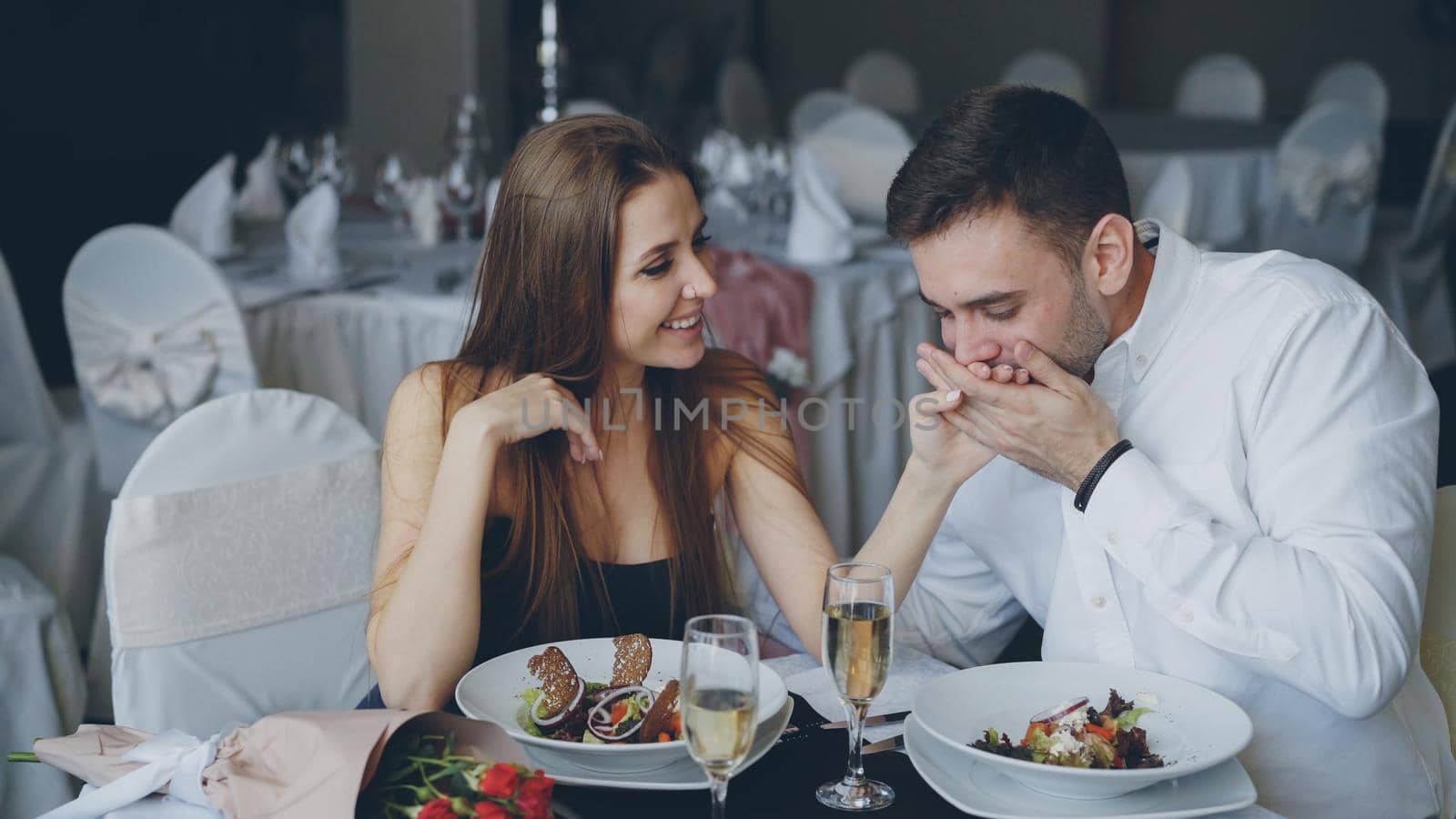 Happy loving couple is holding hands, talking and kissing during romantic dinner in luxurious restaurant. Affectionate relationship, love and fine dining concept.