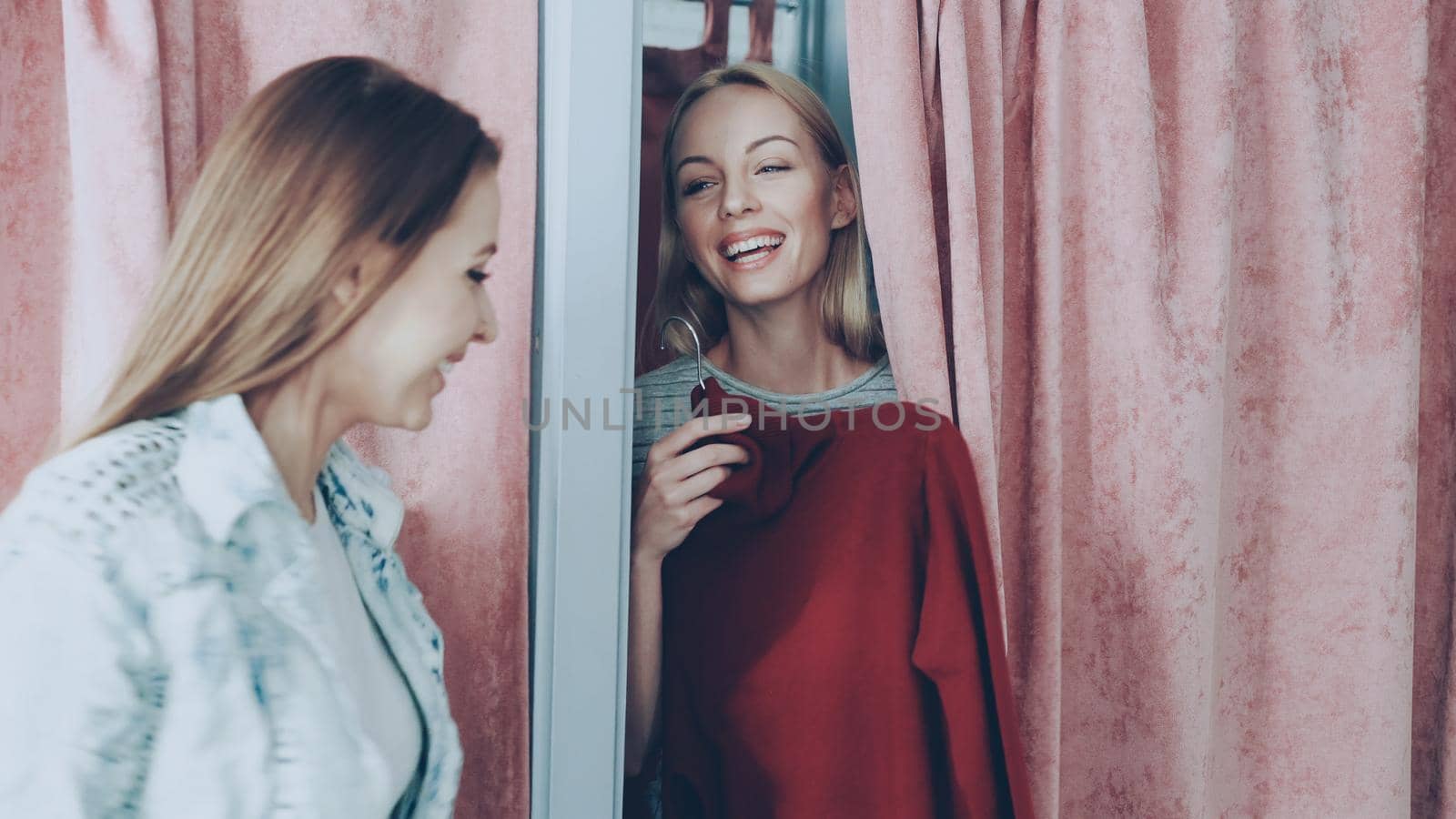 Young blond woman half hidden behind curtain in fitting room in a store asking to fetch her red knitted dress. Female friend bringing her clothes, and she drawing curtain to try it on