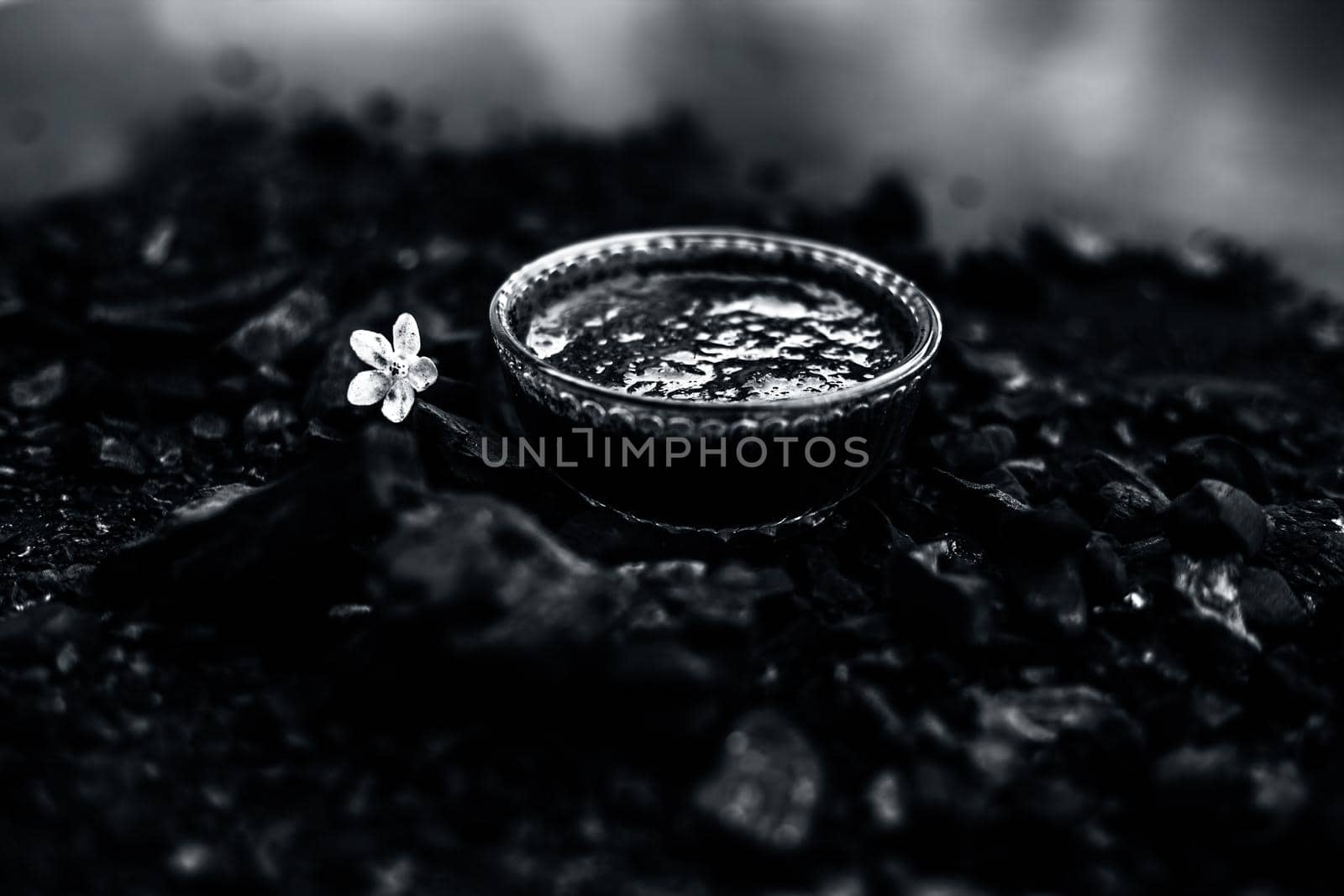 Close up of activated charcoal in a glass bowl on the wooden surface along with some raw powder of charcoal or coal spread on the surface.