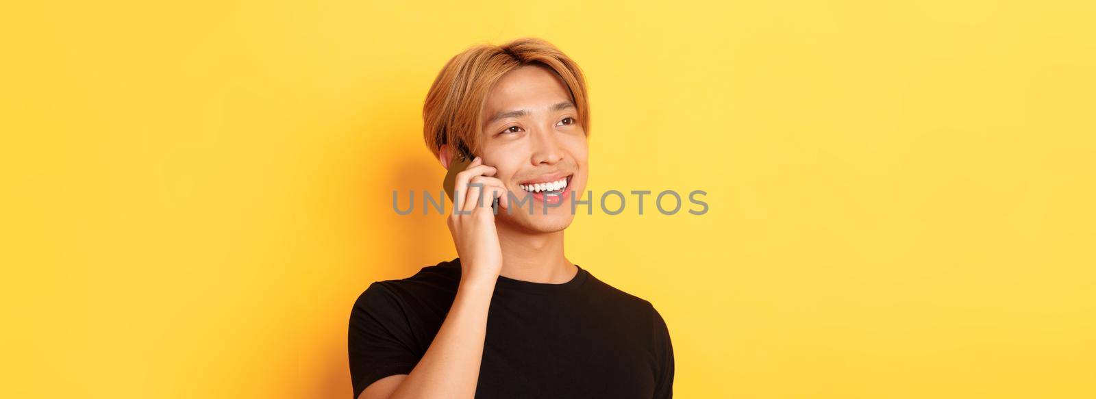 Close-up of handsome carefree asian guy with fair hair, talking on mobile phone and smiling happy, standing yellow background by Benzoix