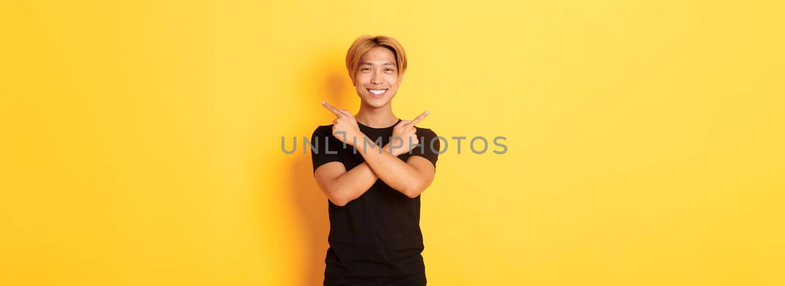 Portrait of handsome determined blond asian guy, student pointing fingers sideways, showing two variants, standing yellow background by Benzoix