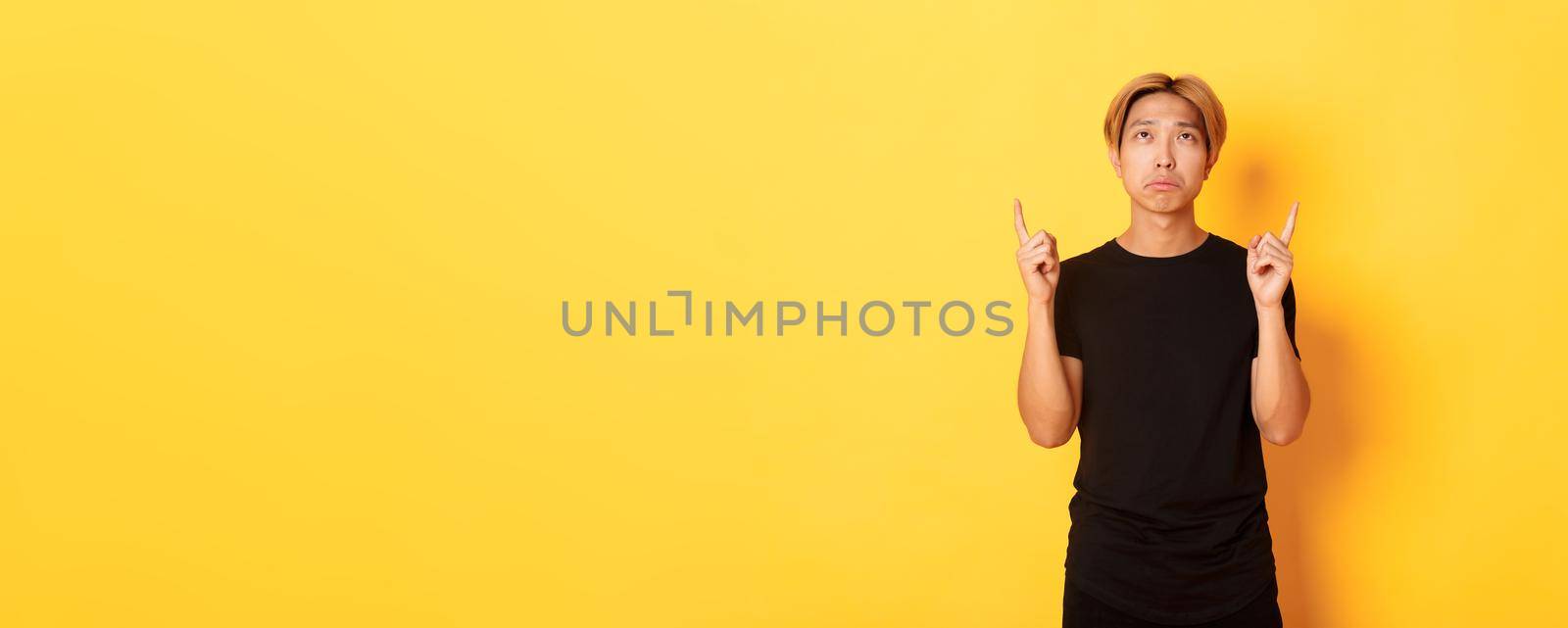 Portrait of gloomy asian blond man, wearing black t-shirt, sulking disappointed and pointing fingers up, yellow background by Benzoix