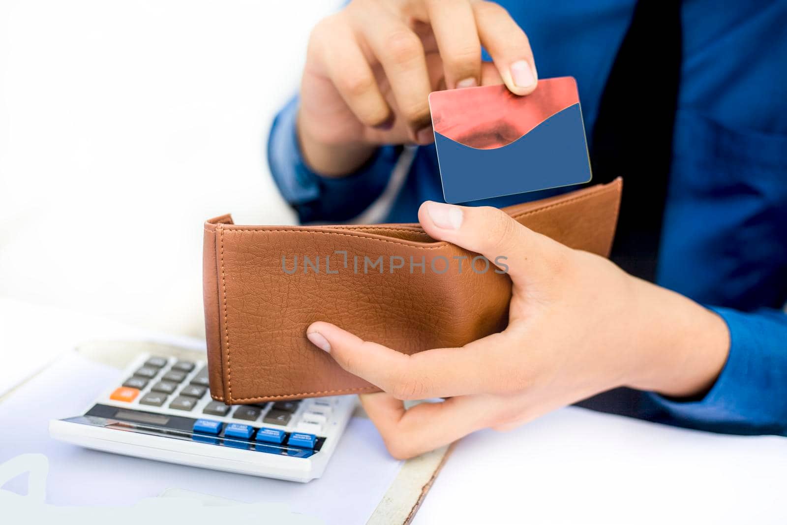 Businessman wearing a dark blue colored shirt with black tie and taking out his credit card from his brown colored wallet isolated on white. by mirzamlk