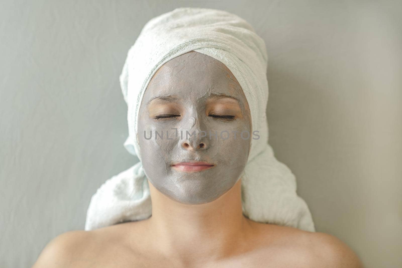 Young woman with clay mask on face and towel on head in form of turban lies with closed eyes and enjoys cosmetic spa treatment for facial skin care. Face close-up, top view.
