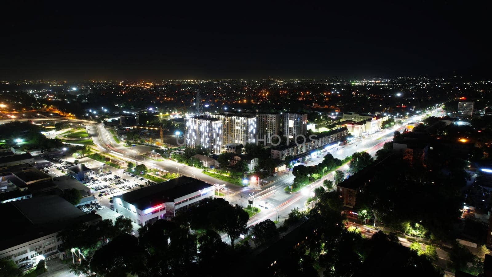 View from the height of the night city of Almaty. Different lights are shining brightly. Cars are driving along the intersection and the bridge. Green trees are growing. High-rise buildings stand out