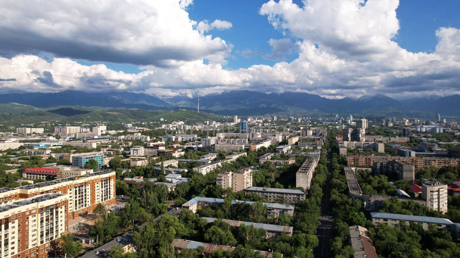 Large dark white clouds over the city of Almaty. Aerial view of the high snowy mountains from the drone. There is a TV tower on the hill. There are many green trees among the houses. A big city