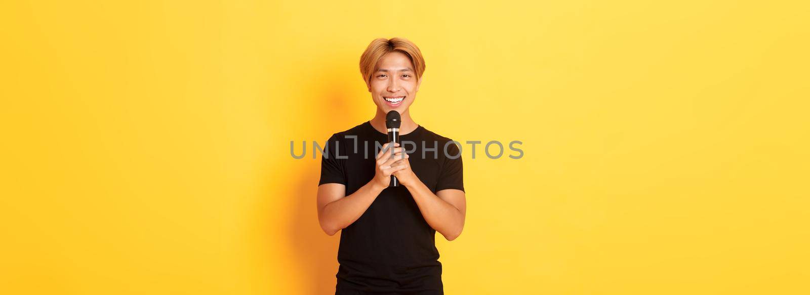 Portrait of charismatic smiling asian man holding microphone and singing karaoke, performing song, standing yellow background.