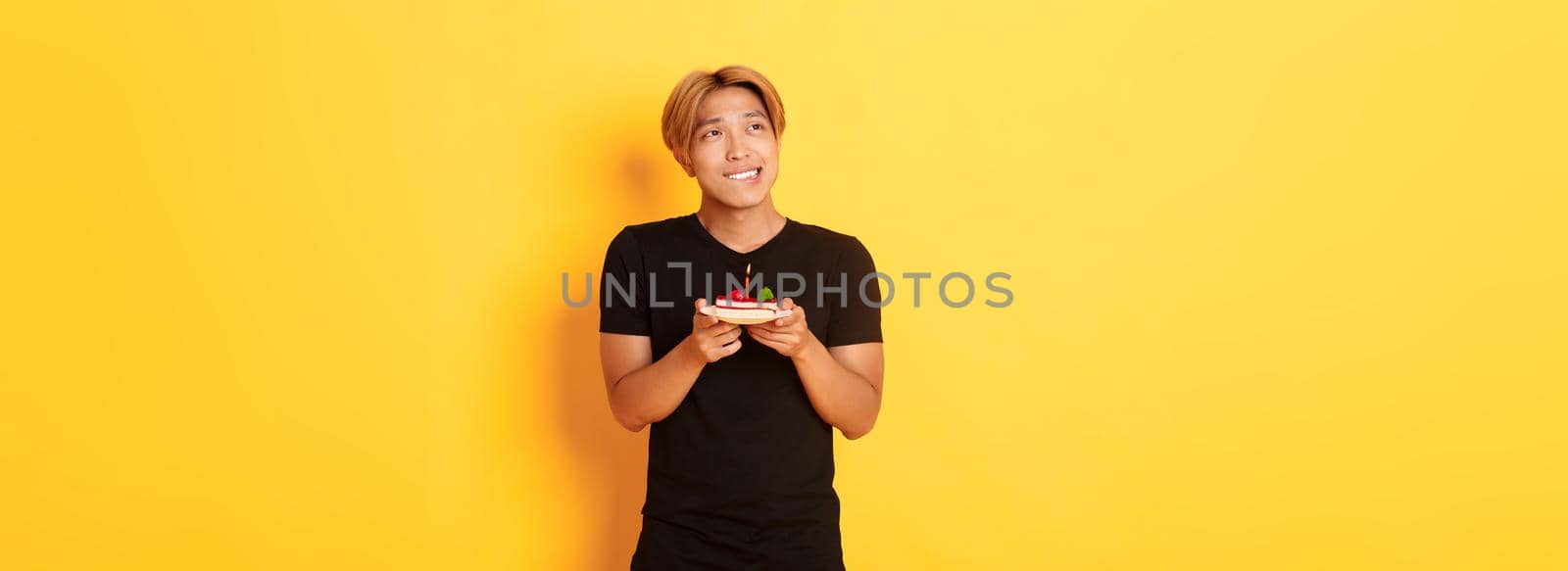 Portrait of handsome dreamy asian guy looking upper left corner and thinking, making wish while celebrating birthday and holding b-day cake, yellow background.