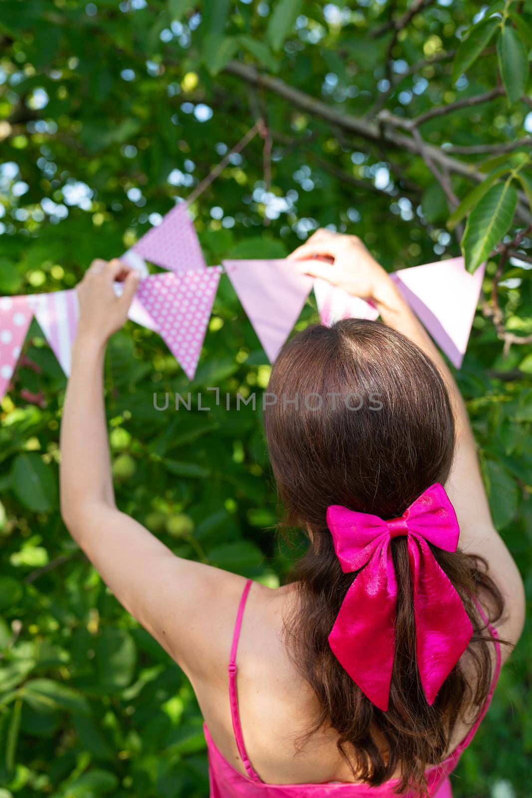 The girl hangs triangular flags of pink color against the backdrop of a green garden. A girl in a pink dress and a bow on her hair. Festive decor - celebration concept. by sfinks