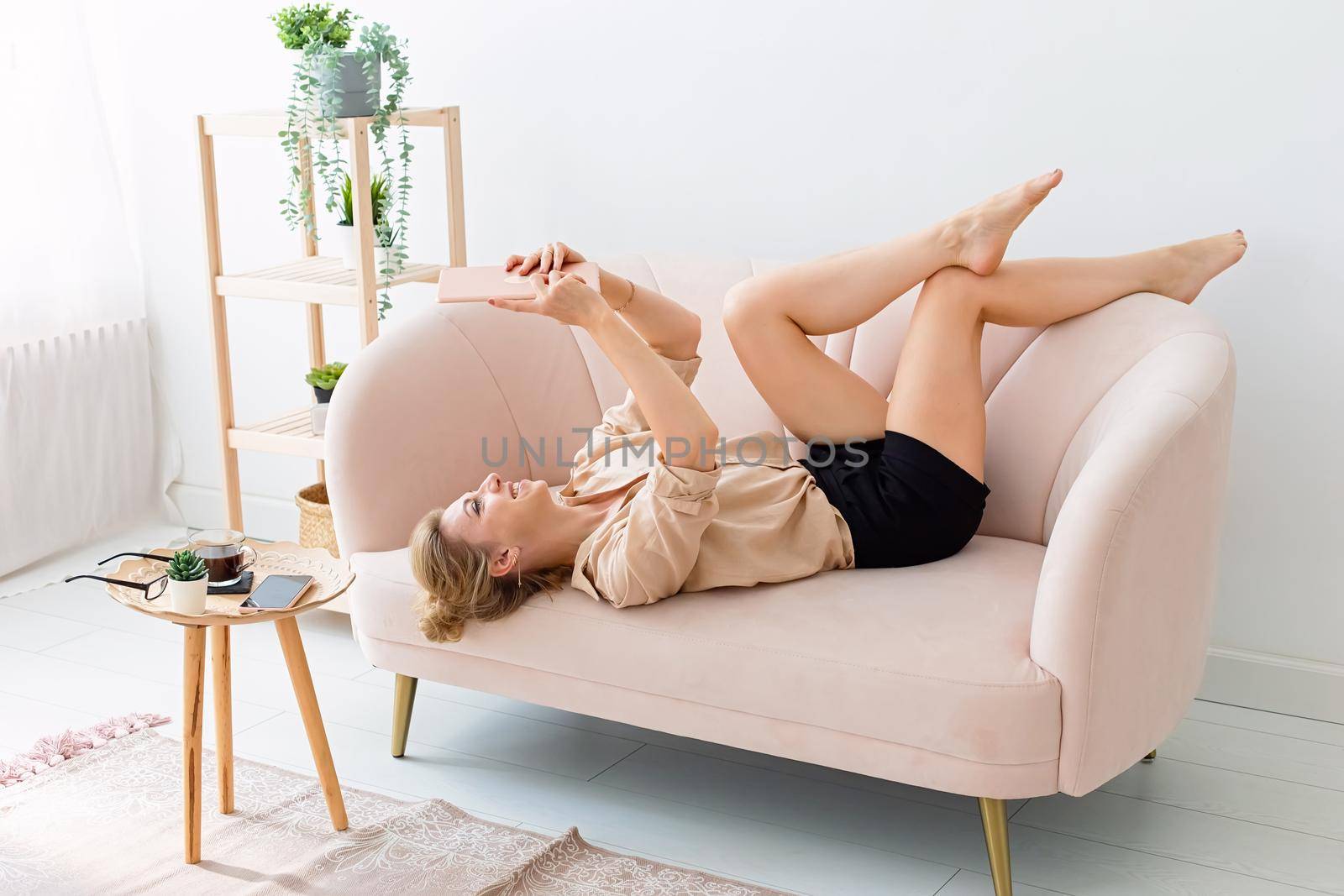 Happy slim blonde 30-40 years old, in a beige shirt and shorts, lies on her back on a beige sofa in a bright living room with plants, legs raised, looks at a digital tablet. Copy space