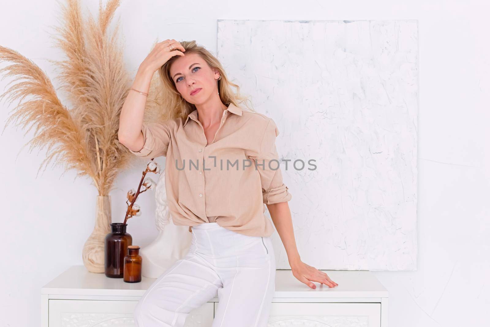 a beautiful slender woman, 30-40 years old, with blond hair, stands in a beige linen shirt and white trousers in the interior of a white room, against a wall and a chest of drawers. Copy space.