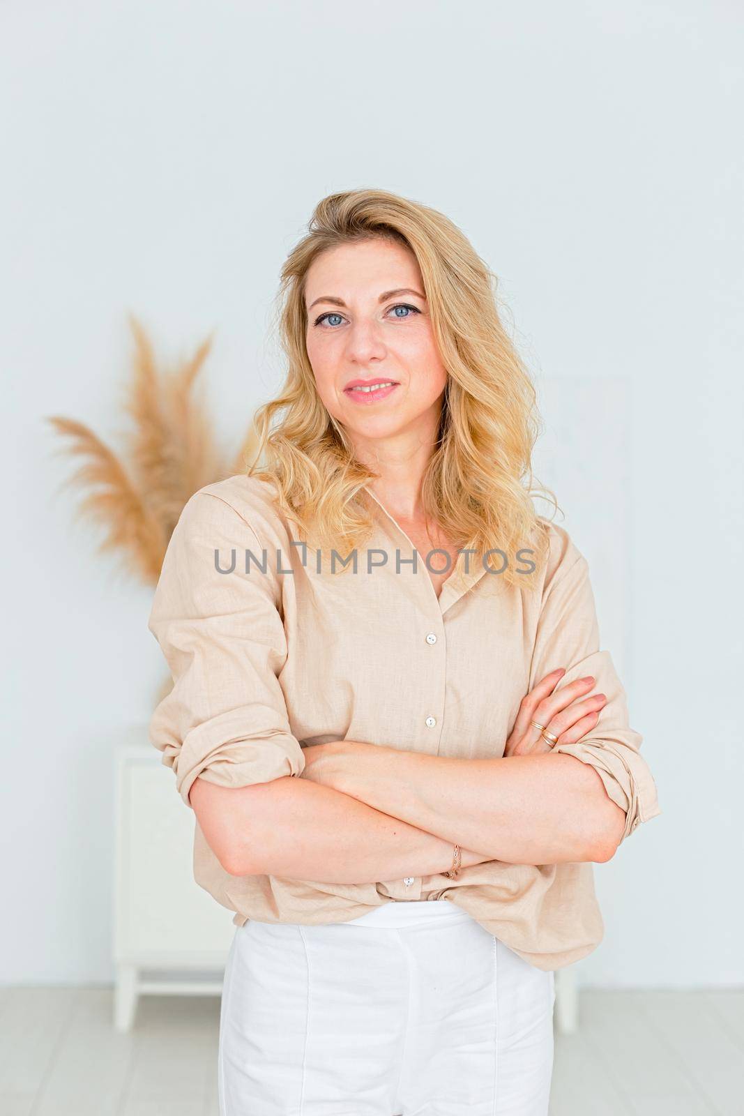 Portrait of a beautiful slender woman, with blond hair, stands in a beige linen shirt and white trousers, in a white room interior. Copy space. Vertical