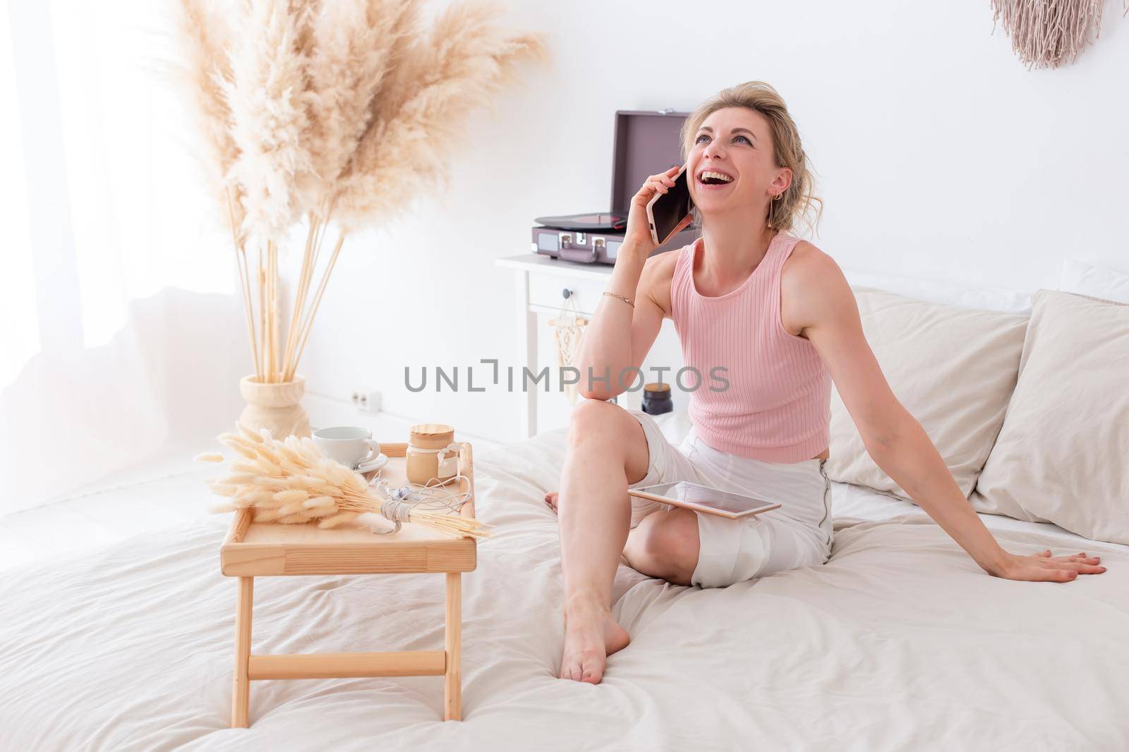 Happy woman with blonde hair, in a pink top and white pants, sitting on the bed in the morning, in a bright living room interior, talking on the smartphone and holding a digital tablet on her knees. Copy space