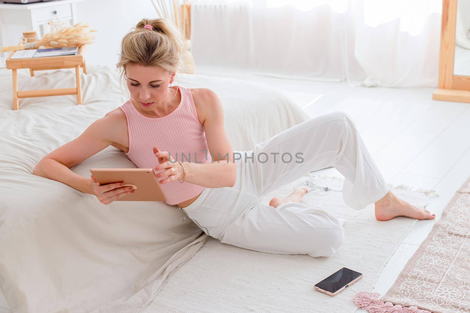 A beautiful slender woman with blond hair, a pink top and white pants, sits on the floor in the morning, near a white bed, watches a digital tablet, a smartphone lies nearby
