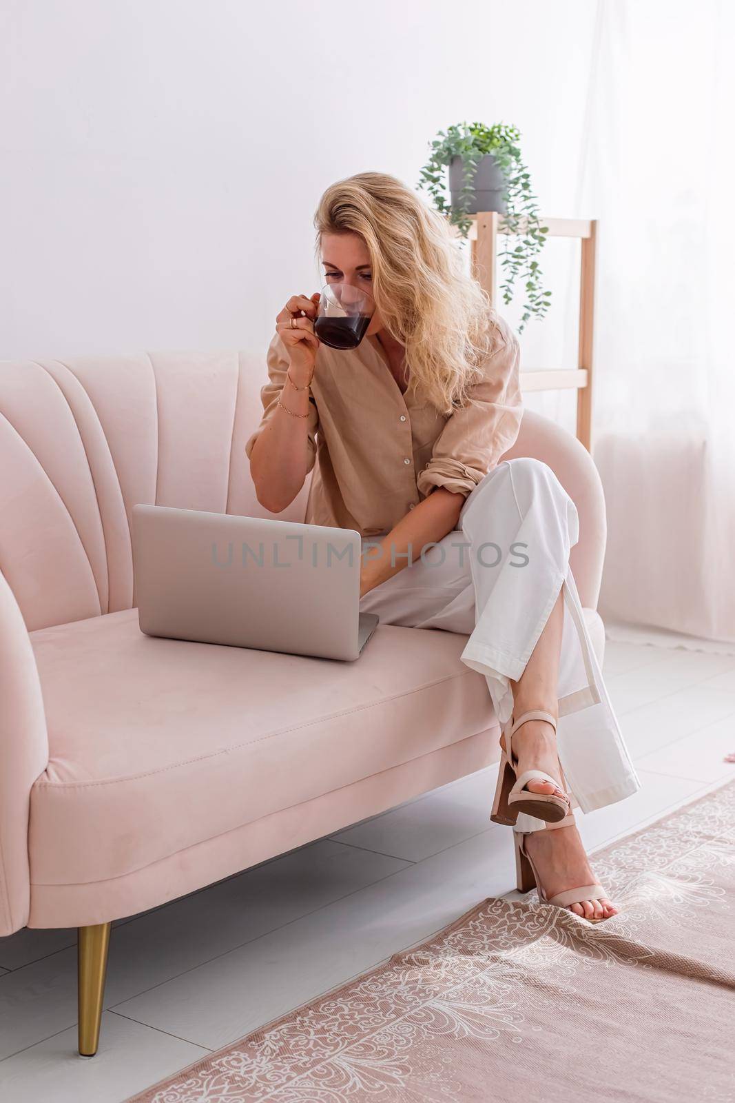 One stylish woman in light clothes sitting on light pink sofa, working on laptop, drink a cup of coffee. Vertical. Copy space