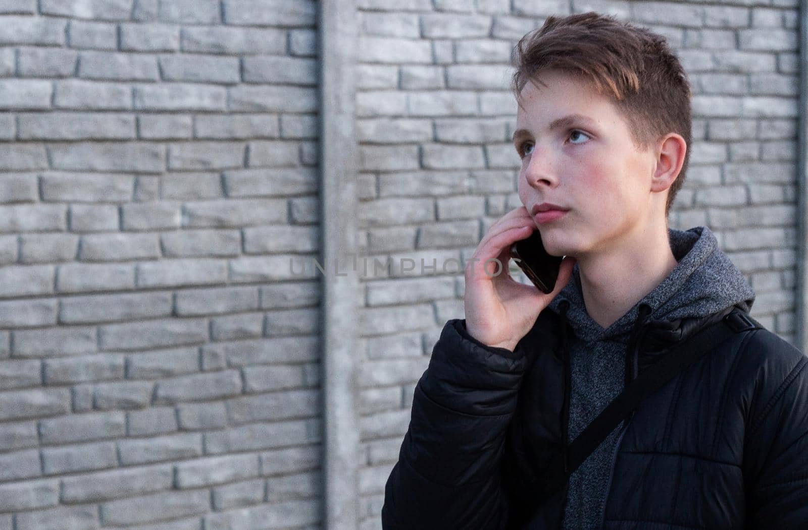 A pensive boy in a gray sweatshirt and black jacket stands in the street and holds a smartphone in his hand. Portrait of an attractive 15-year-old boy with a phone in his hand against a wall..