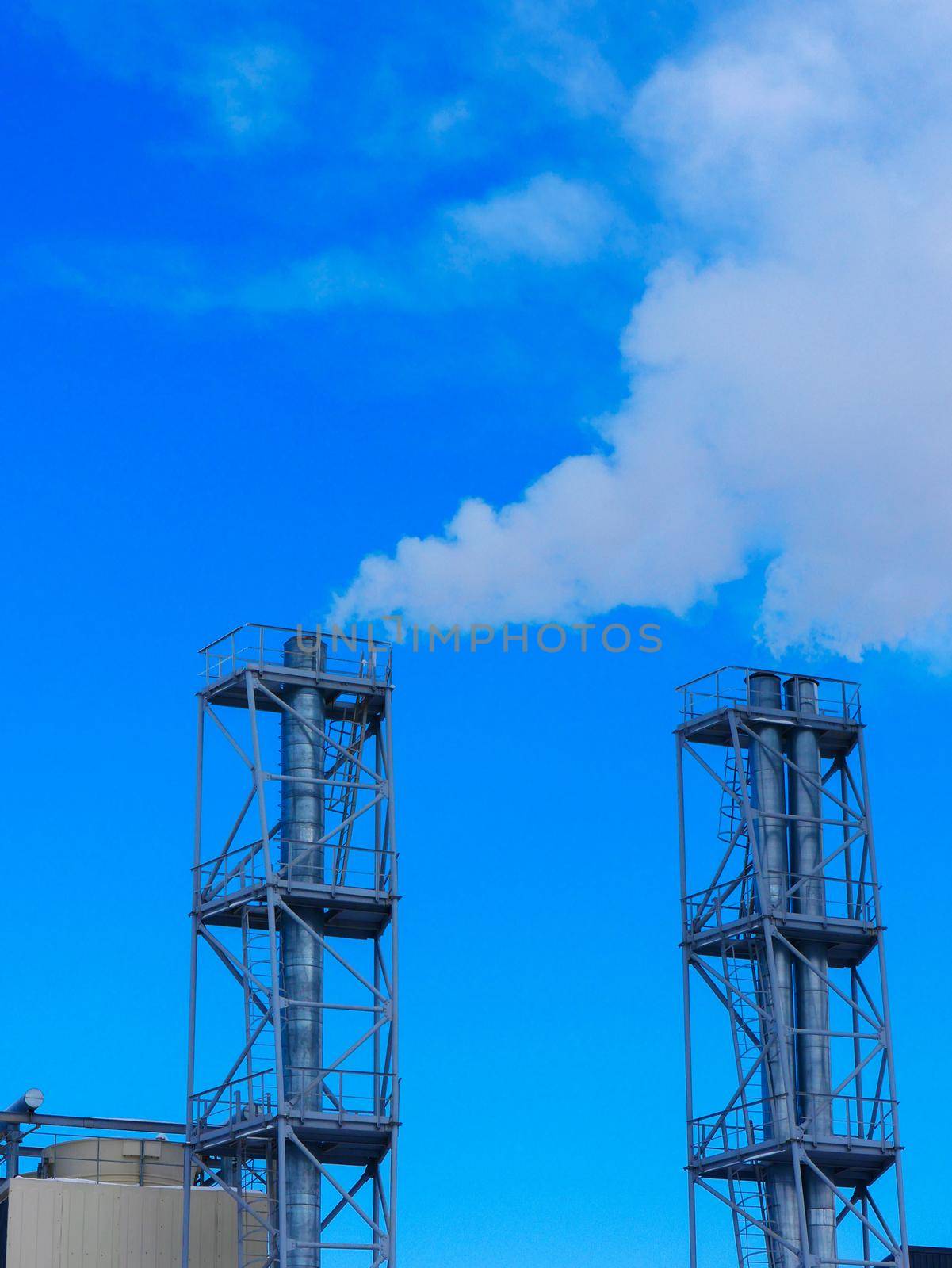 Two chimneys of a heating plant against the background of a bright blue sky and smoke coming out of them.