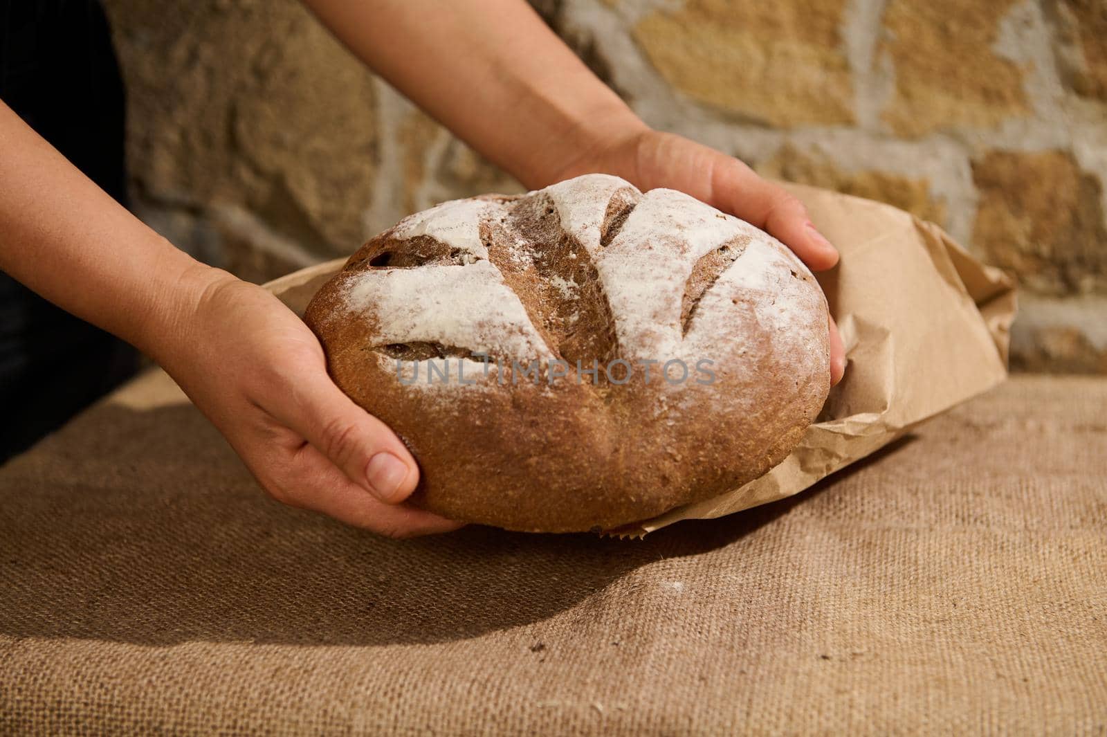 Fresh baked homemade whole grain rye bread in the hands of a housewife, baker, above a table with burlap tablecloth by artgf