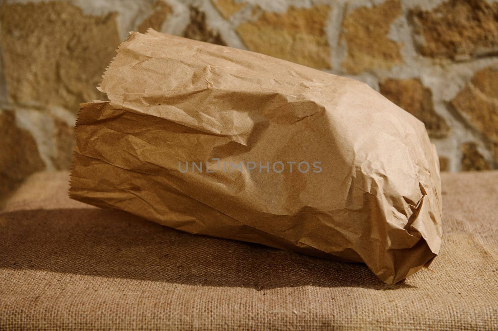 Close-up. Selective focus on an eco-friendly recyclable craft paper bag, with healthy wholegrain wheat bread inside, on a table with a burlap tablecloth against stone wall background. Copy ad space