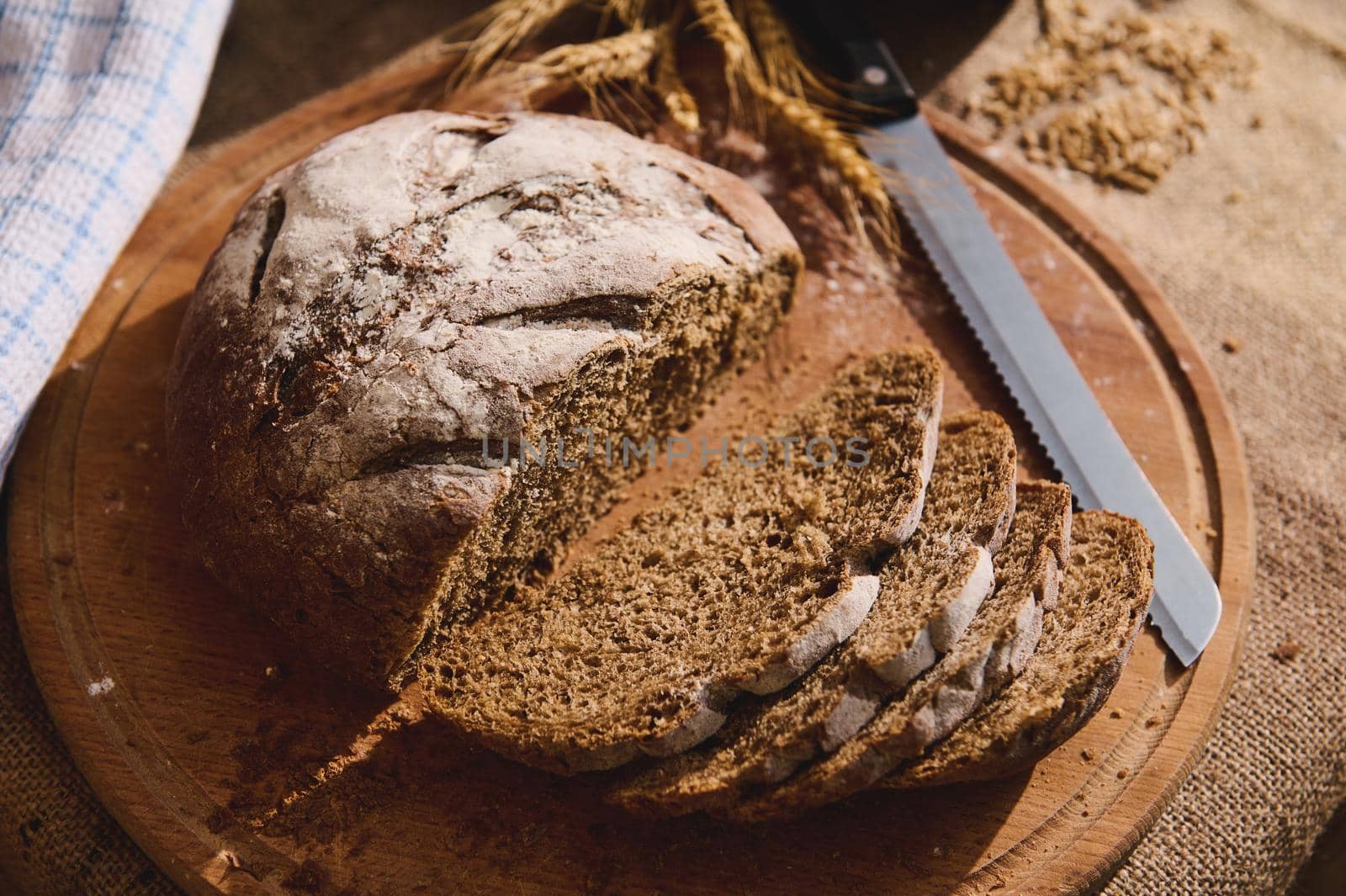 View from above of slices of freshly baked whole grain sourdough bread on a wooden board on a table covered with a burlap tablecloth, wheat ears and scattered cereal grains on a blurred background