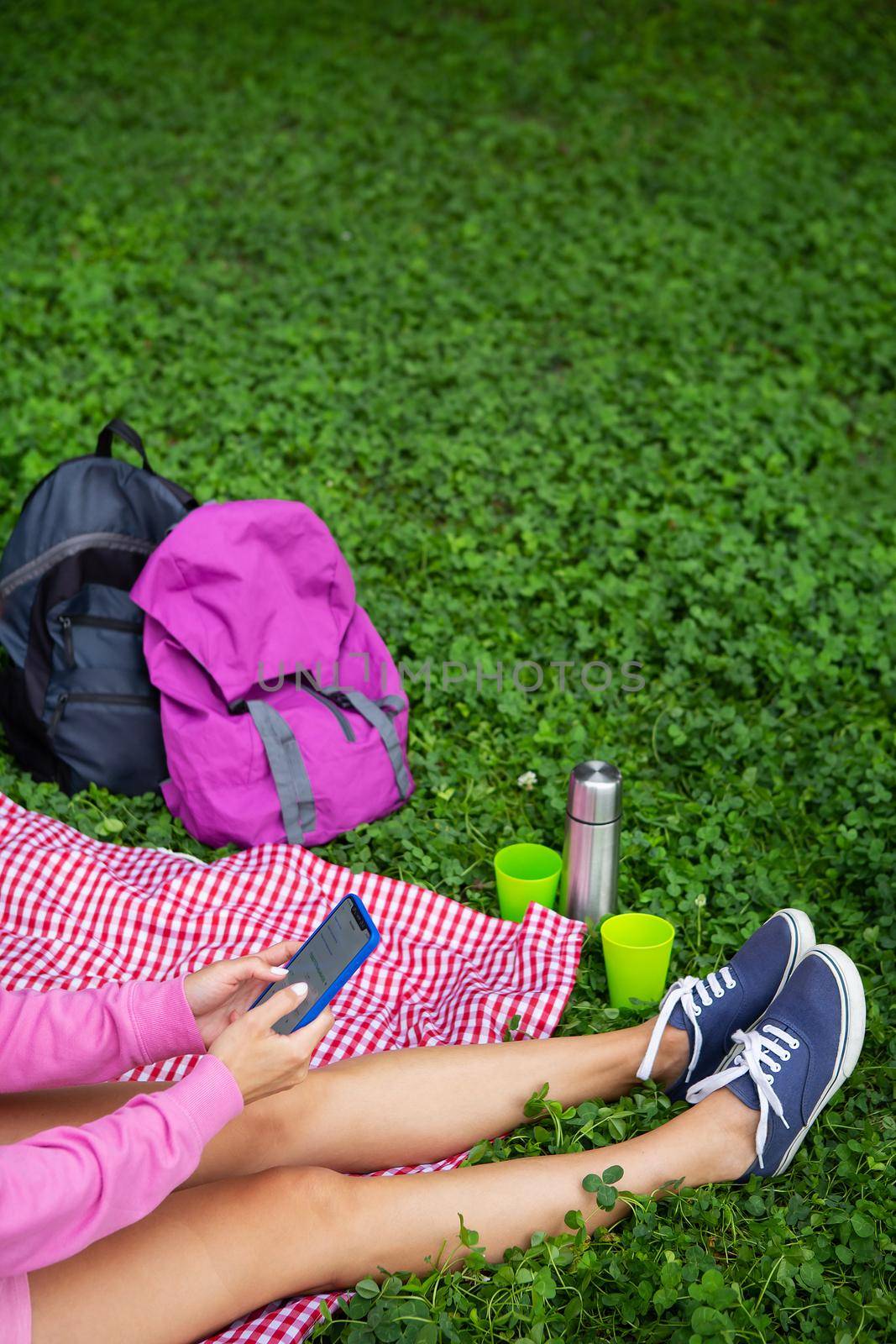 Top view of a young female student sitting on a plaid blanket and green grass in the park. Break in the open air, drink coffee, tea, sit on the phone. by sfinks
