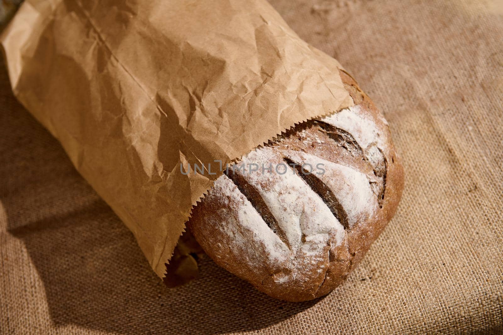 Still life with an artisanal whole grain, healthy rye sourdough bread, in a recyclable eco paper bag on the rustic table with sackcloth tablecloth against a brick and stone wall background. Top view