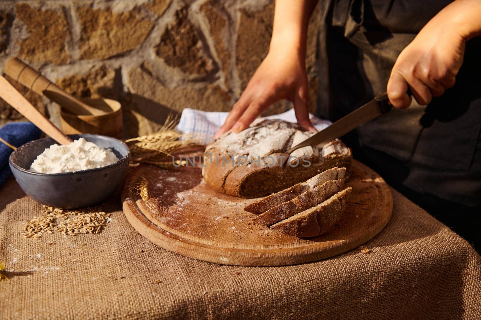 Baker cuts a loaf of rye bread on wooden board. White flour and wheat spikelets on table, covered with burlap tablecloth by artgf