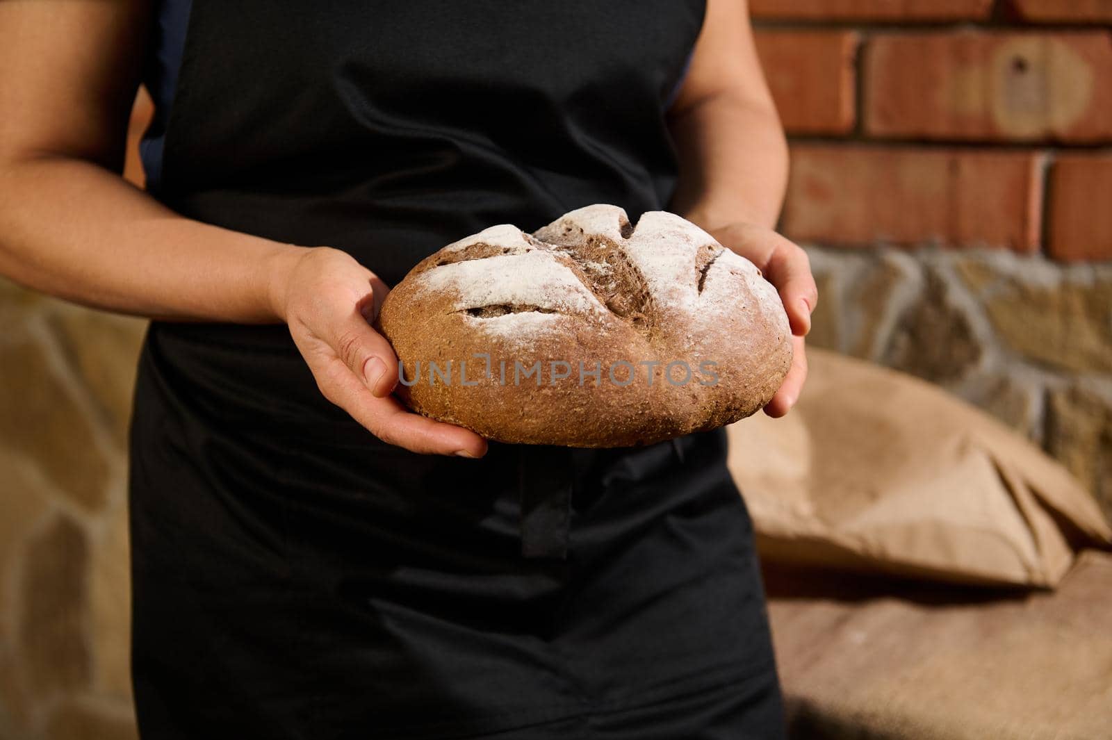 Cropped view of a housewife, a female baker in black chef's apron holding a loaf of fresh baked warm whole grain wheat bread in her hands, standing against a brick wall background. Baking concept