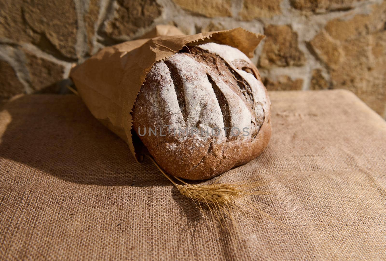 Wholesome whole grain rye bread in a craft eco-paper bag and ears of wheat on a table covered with a burlap tablecloth. Concept of baking healthy homemade bread. Selective focus. Copy ad space