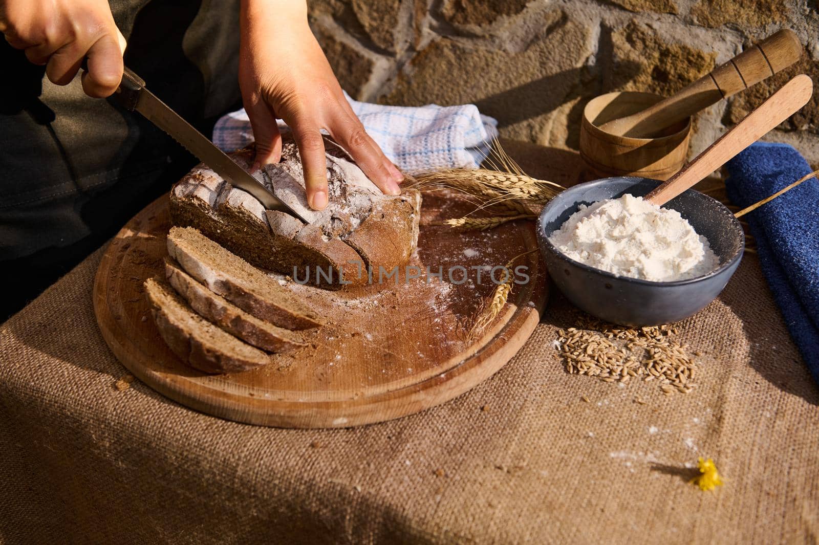 Selective focus on the hands of a baker using knife, slices a loaf of a whole grain sourdough rye bread on a wooden cutting board. White flour and wheat spikelets on the table. Artisanal rustic bakery