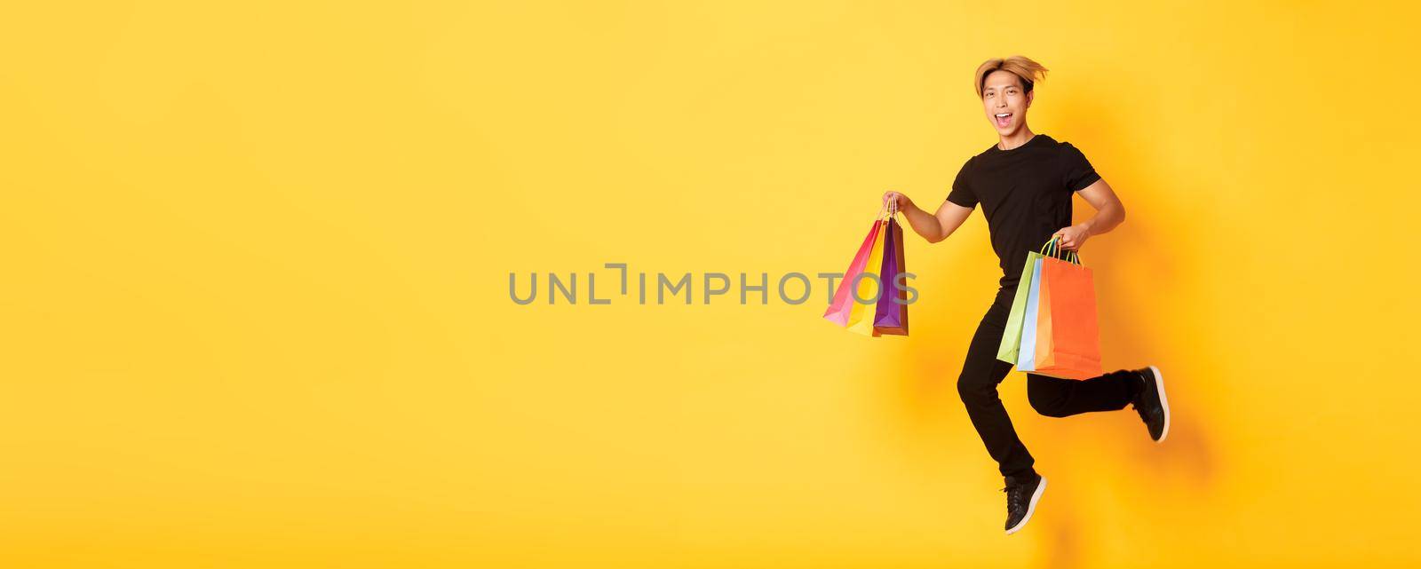 Full-length of happy attractive asian man jumping from happiness and carry shopping bags, standing yellow background.
