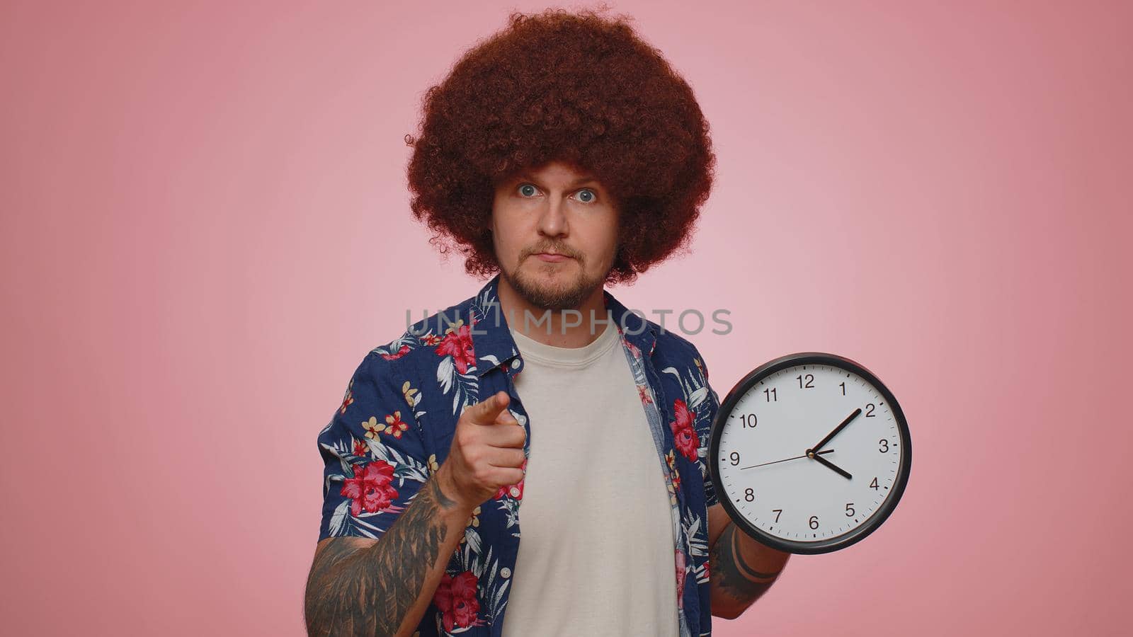 It is your time. Portrait of bearded young man in shirt showing time on clock watch, ok, thumb up, approve, pointing finger at camera. Adult guy indoors studio shot isolated alone on pink background