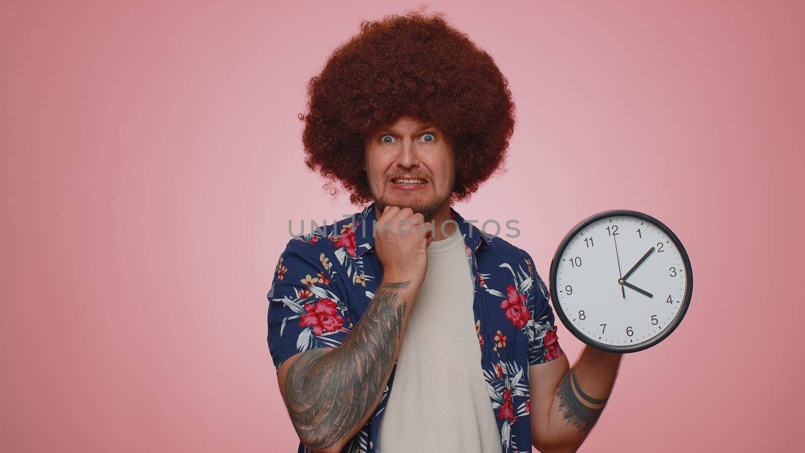 Confused young man with anxiety checking time on clock, running late to work, being in delay, deadline on pink studio background. Serious guy looking at hour and minutes, worrying to be punctual