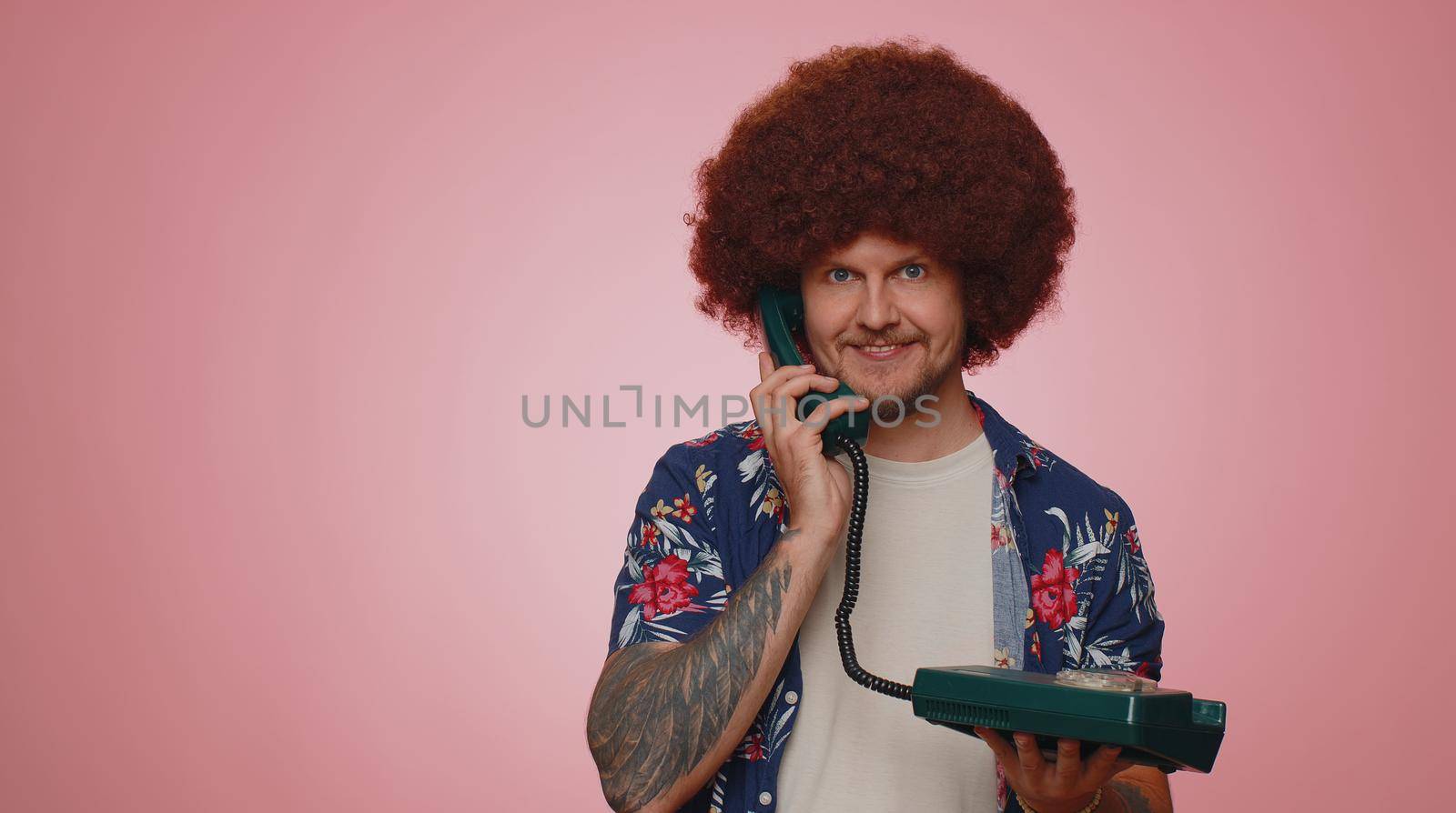 Hey you, call me back. Cheerful man secretary in blue shirt talking on wired vintage telephone of 80s, says hey you call me back. Young adult guy posing isolated on pink studio wall background