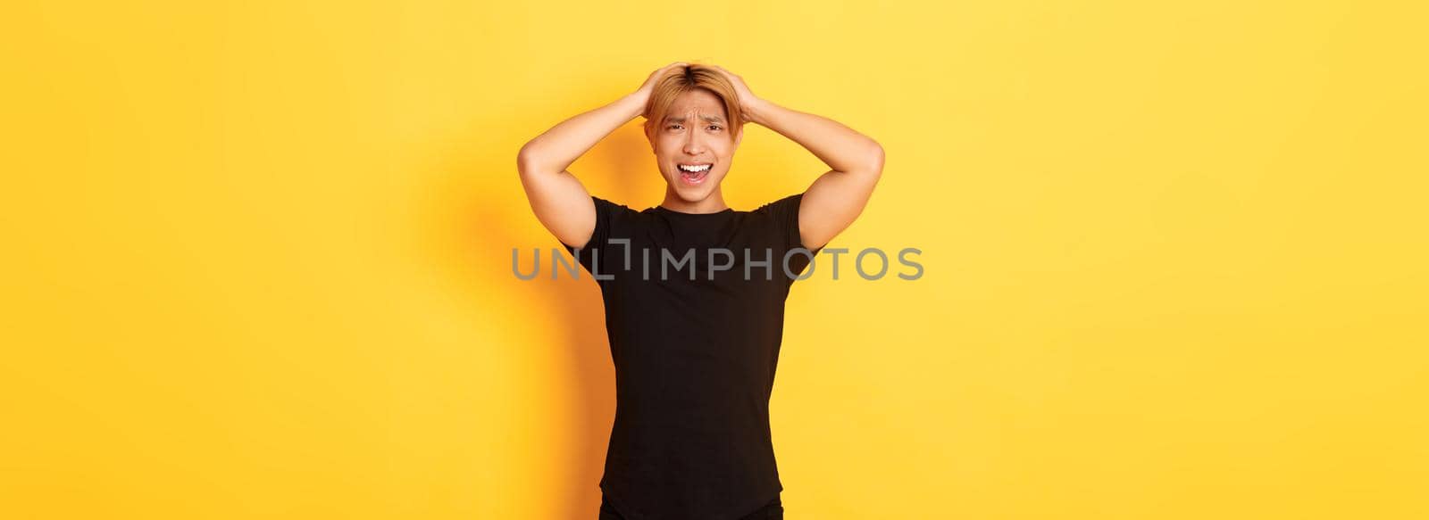 Portrait of young asian guy panicking, looking anxious and upset, holding hands on head and grimacing, standing over yellow background by Benzoix