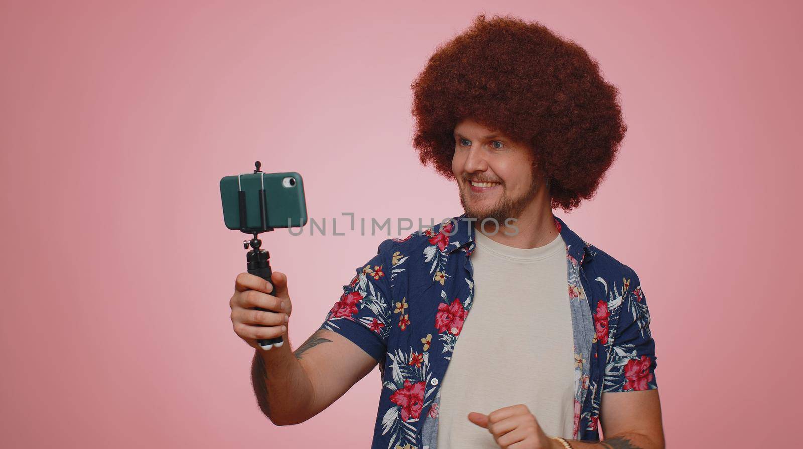 Tourist man in shirt traveler blogger taking selfie on mobile phone selfie stick communicating video call online with subscribers. Young adult guy boy 30s isolated alone on pink studio wall background