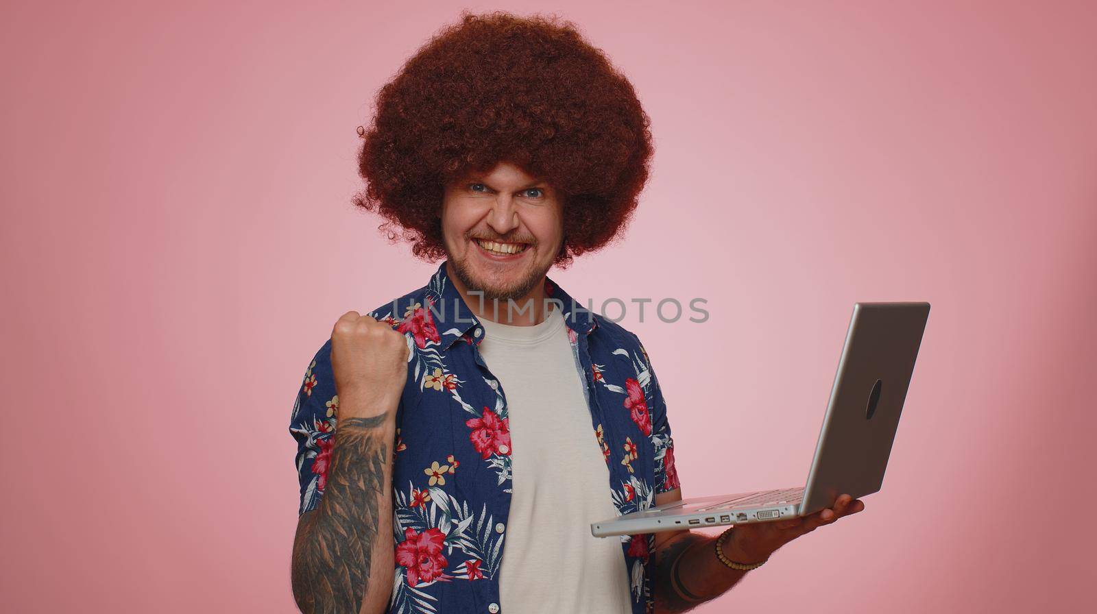Excited happy tourist man typing on laptop computer, working on project, enjoying results, winning lottery, celebrating success, online shopping. Young bearded guy boy isolated on blue background