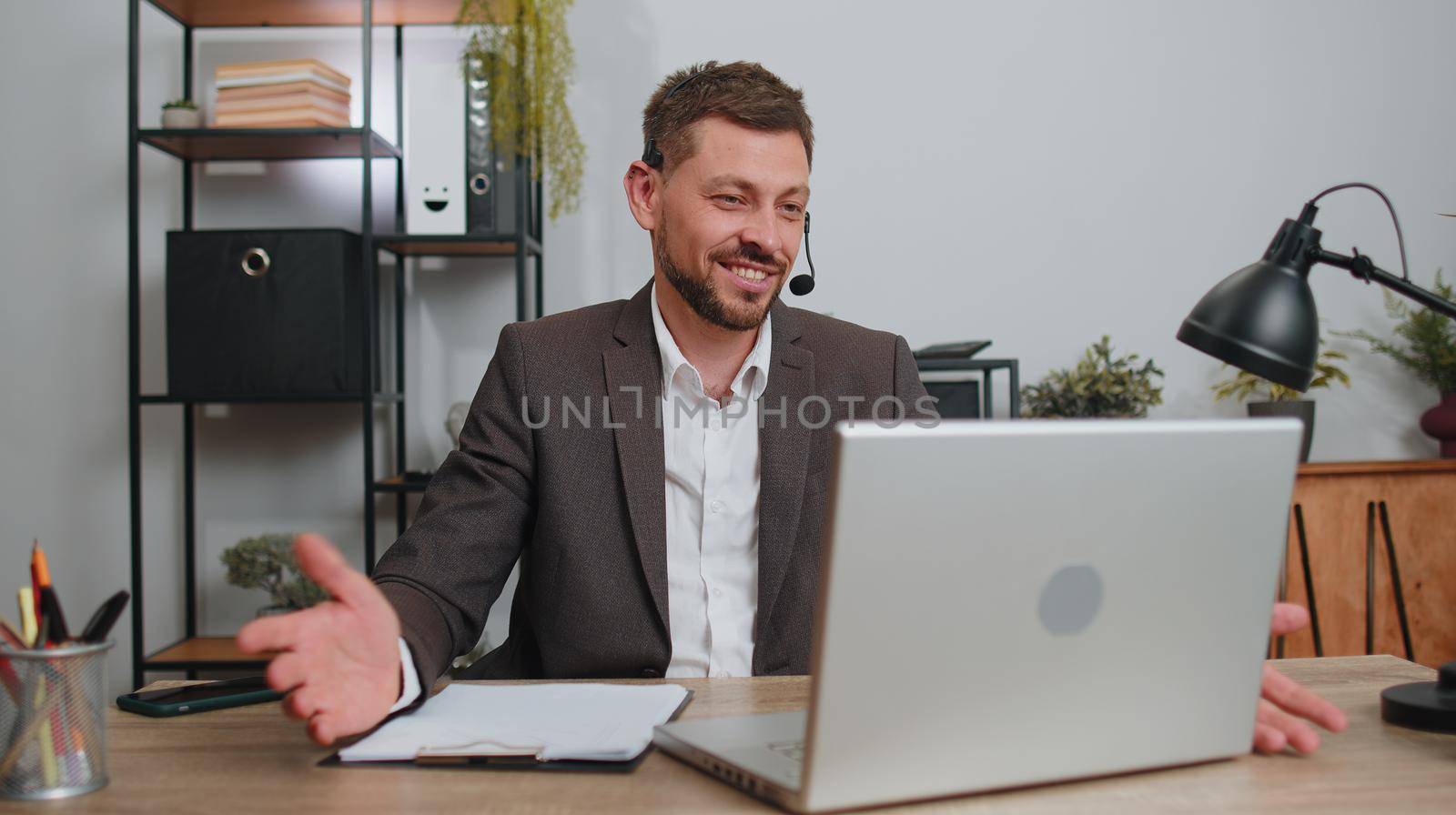 Businessman working on laptop computer wearing headset, freelance worker call center or support service operator helpline, having pleasant talk with client or colleague communication support at office
