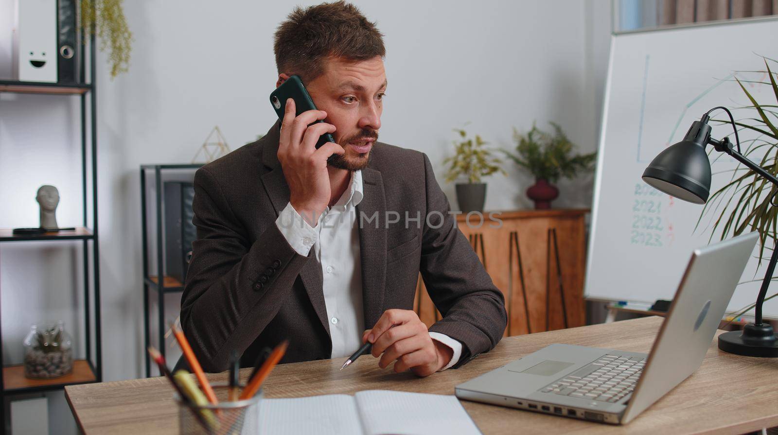 Conflict, quarrel, negotiation. Angry businessman ceo in formal wear suit making phone call at home office desk. Concentrated upset mad male freelancer man talking smartphone conversation indoors