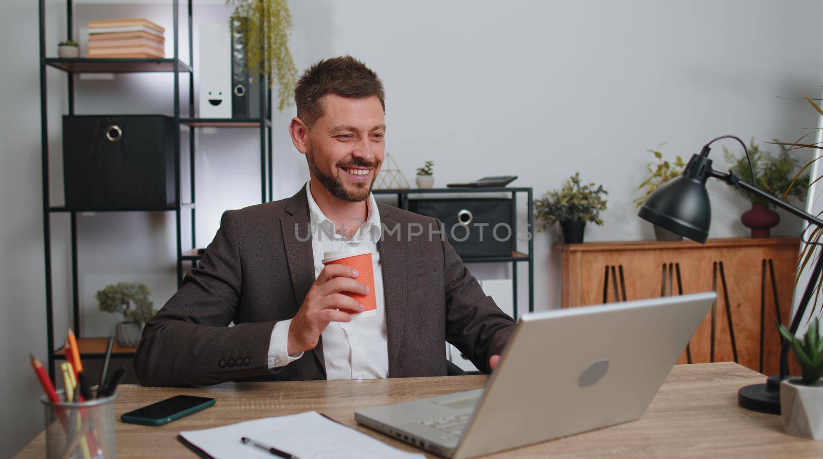 Businessman freelancer enters office start working on laptop computer at desk and drinking morning coffee. Man works on notebook, sends messages makes online purchases watching social media movies