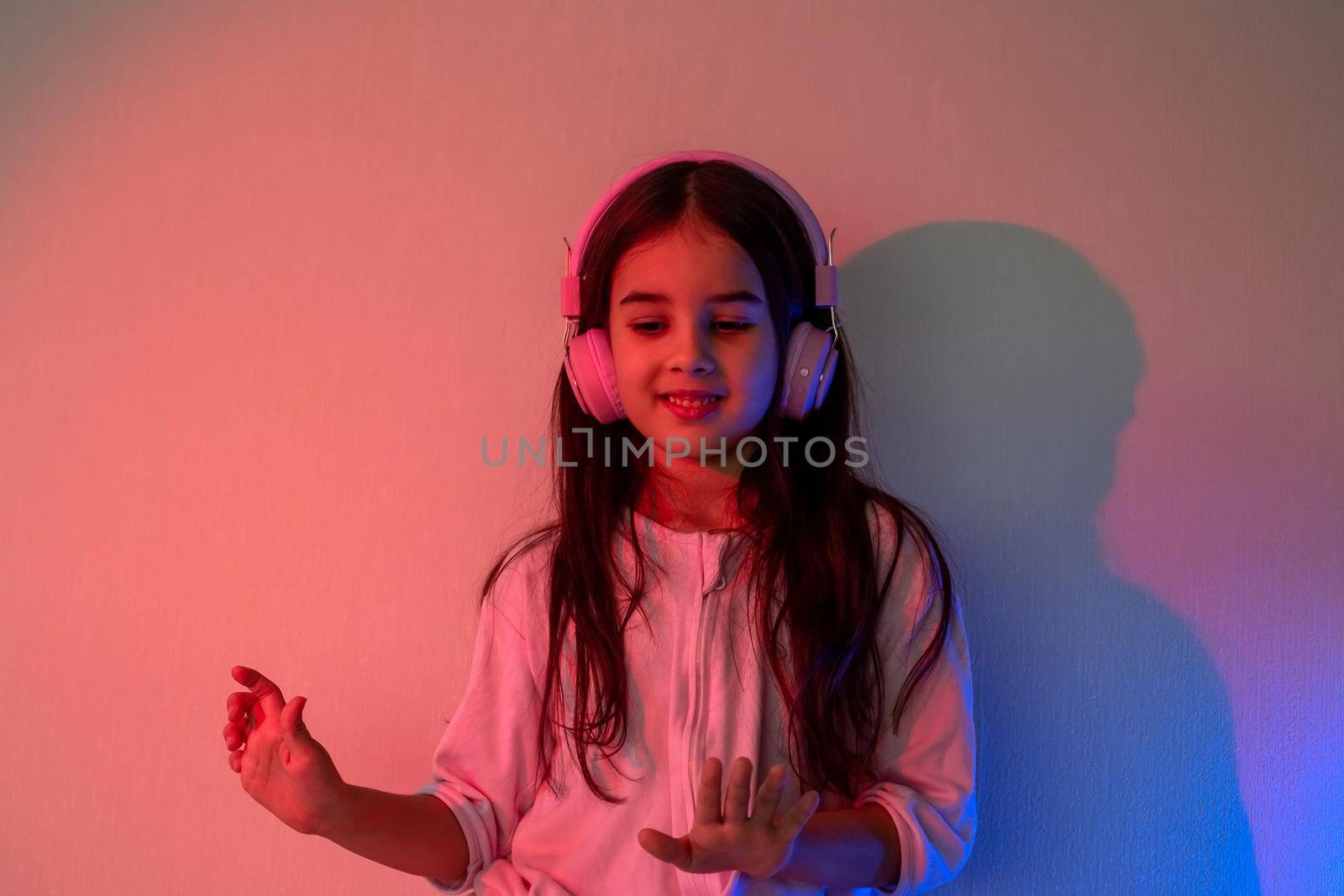 Little girl in pink headphones and cozy clothes dances to music in neon pink blue light