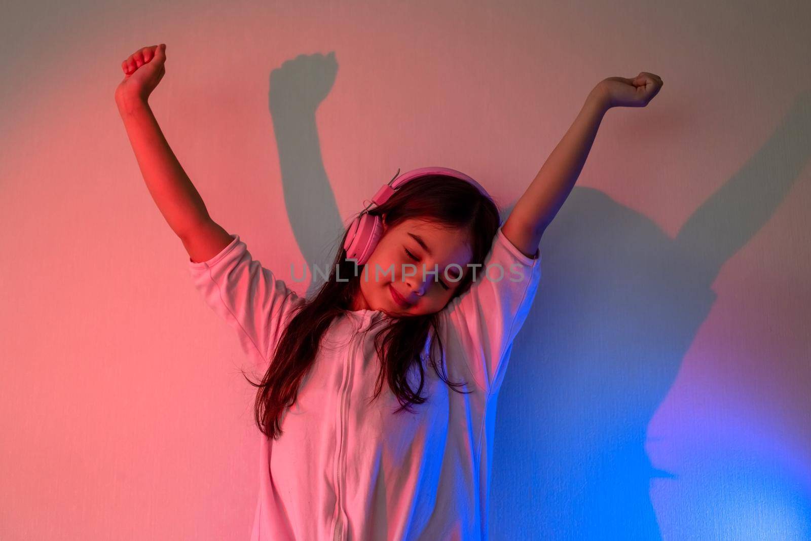 Dancing little girl in pink headphones with loose dark hair in neon pink and blue light against the wall, raised her hands up and closed her eyes