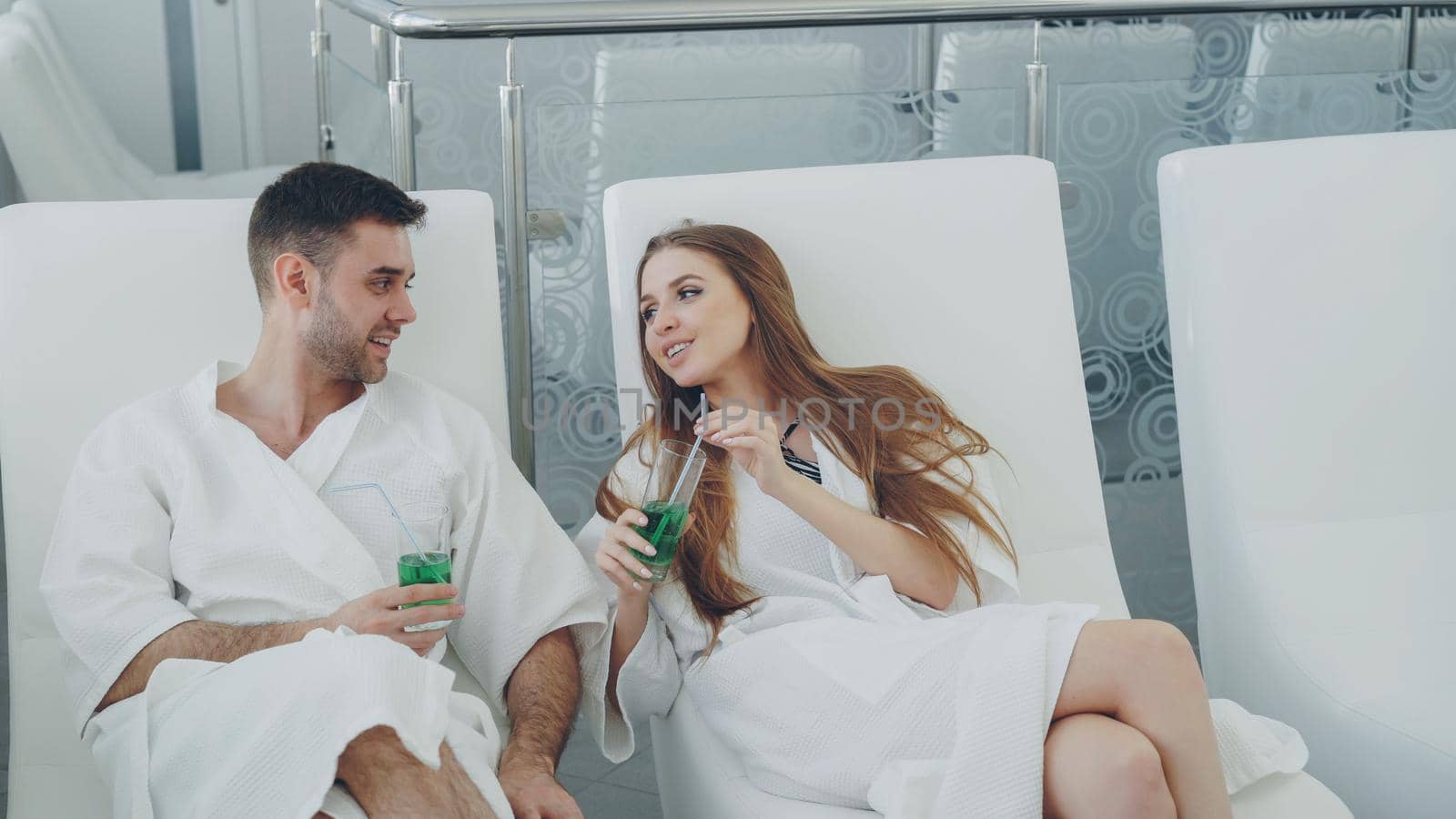 Young lovers in bathrobes are relaxing in chairs in spa salon, drinking cocktails and talking. Wellness, romantic relationship and happiness concept.