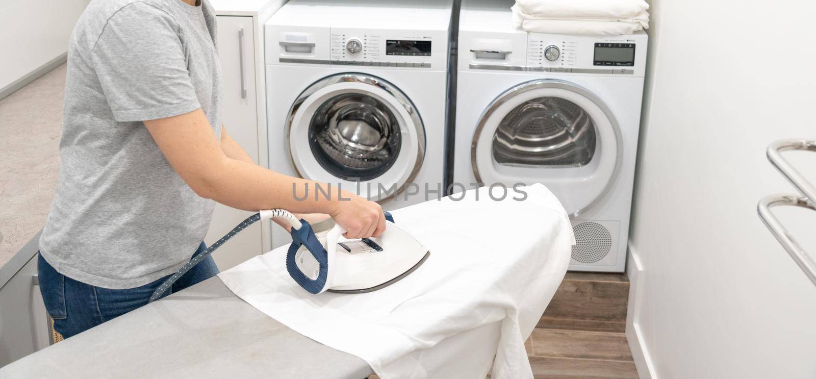 Woman ironing white shirt on board in laundry room with washing machine on background by Mariakray