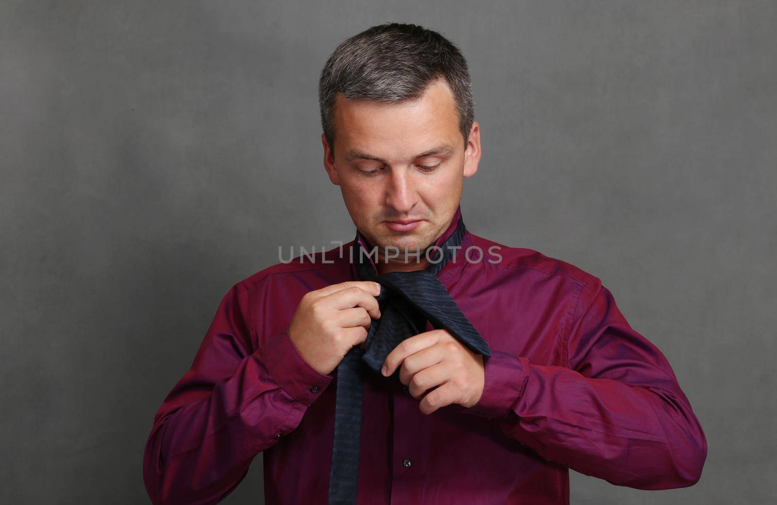 A man in a red shirt on a grey background with a tie..A fashionable and handsome man is trying to tie a tie.