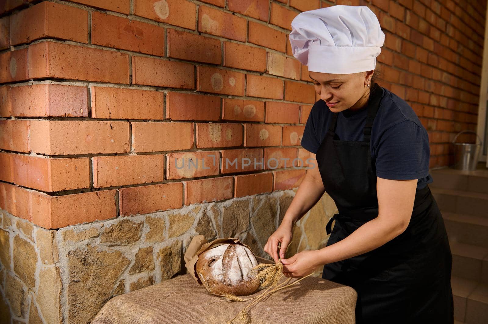 Female baker chef in a black apron and white cap, places wheat spikelets next to freshly baked whole grain bread on a table covered with burlap tablecloth, putting it up for sale with fresh pastries