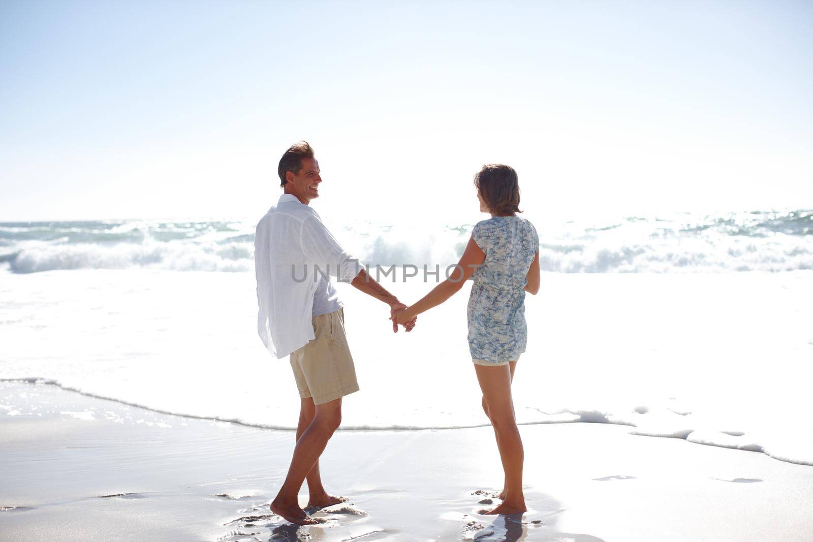 Enjoying the romance of the ocean. Rearview of a happy mature couple walking hand in hand on the beach. by YuriArcurs