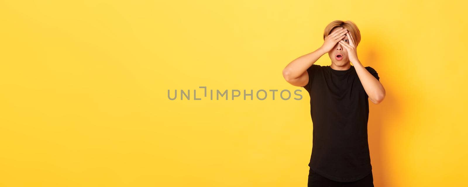 Portrait of embarrassed asian guy with blond hair, gasping startled and shut eyes, peeking through fingers, yellow background.