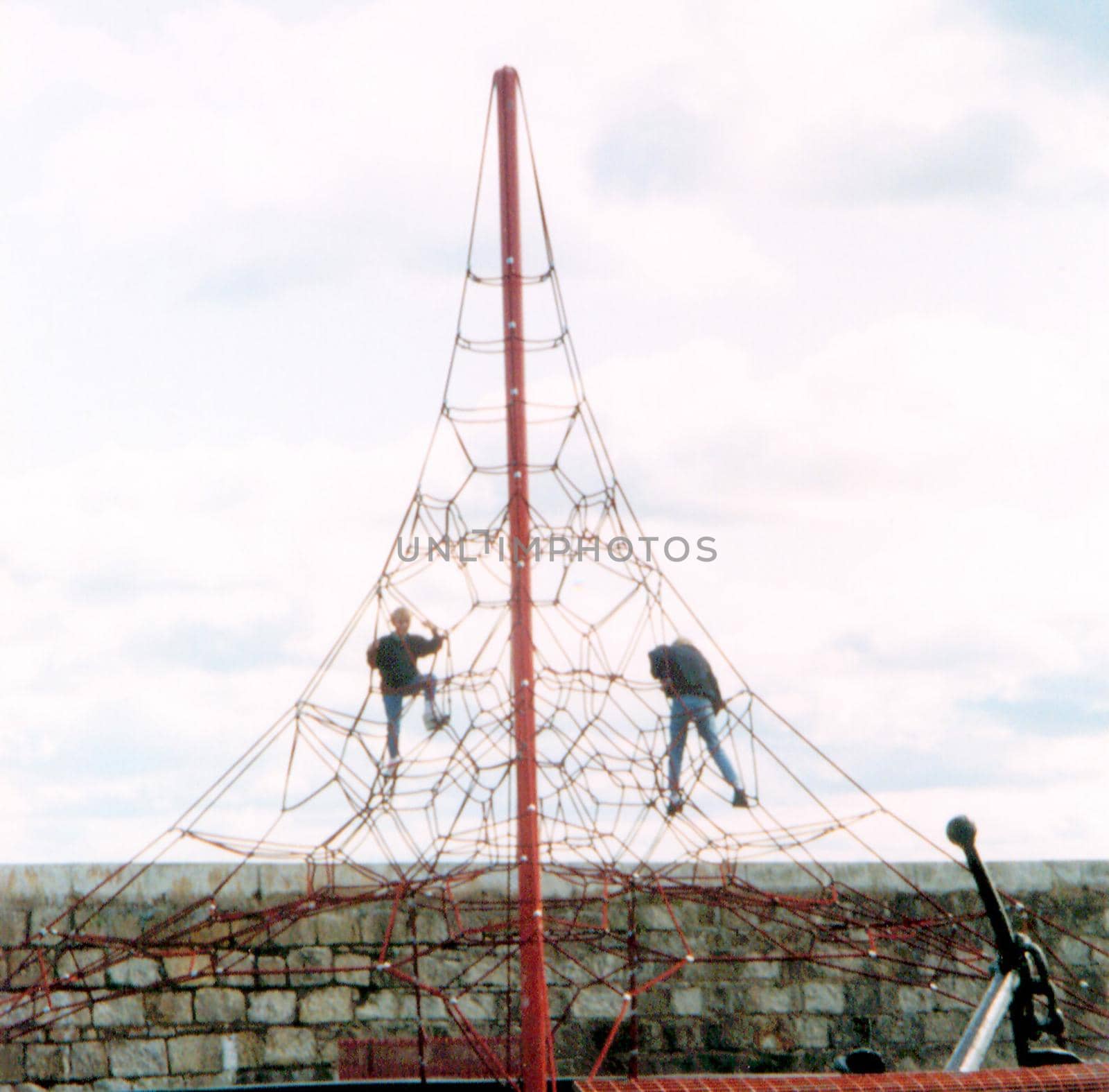 Two young men climbing on a exterior tall play frame in front of an old stone wall an overcast sky