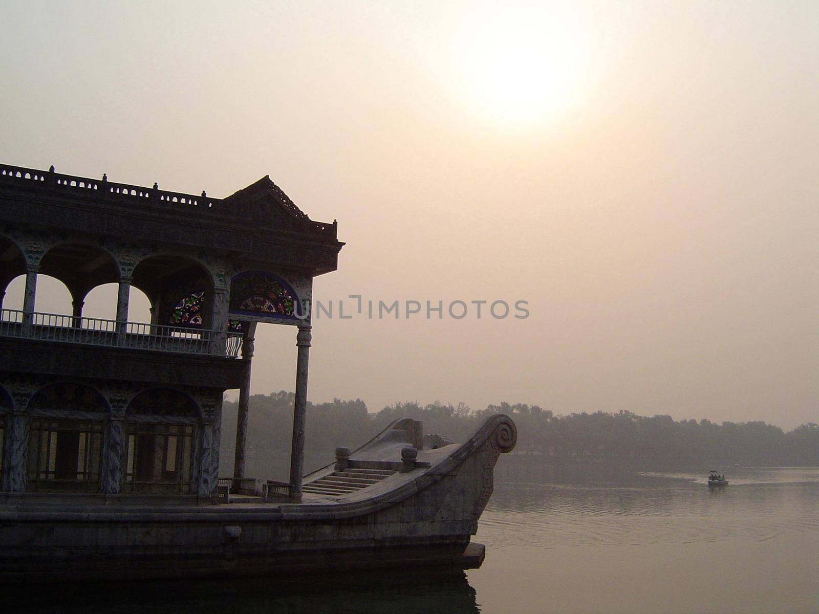 Detail of House Boat with Ornate Carvings Floating on Calm River with Small Boat and Shoreline in Background Under Hazy Sky with Obscured Sun